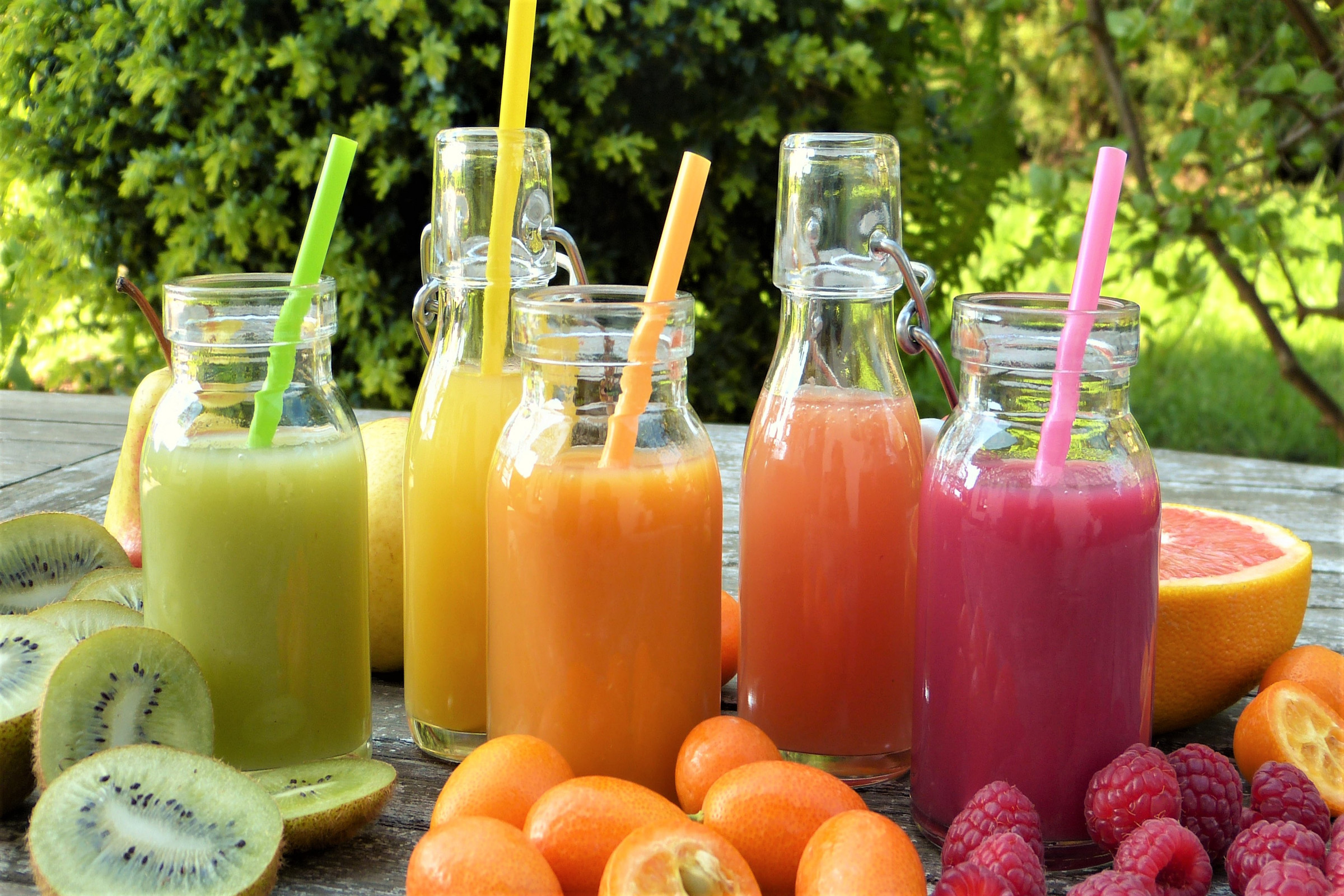 Cinco copos com sucos de cores e sabores diferentes e frutas atrás dos copos